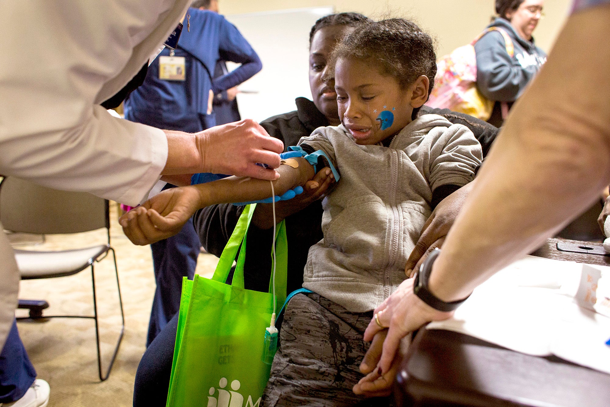 A boy sits on his mothers lap and cries as his blood is drawn by a person in a white lab coat who is mostly out of the frame.