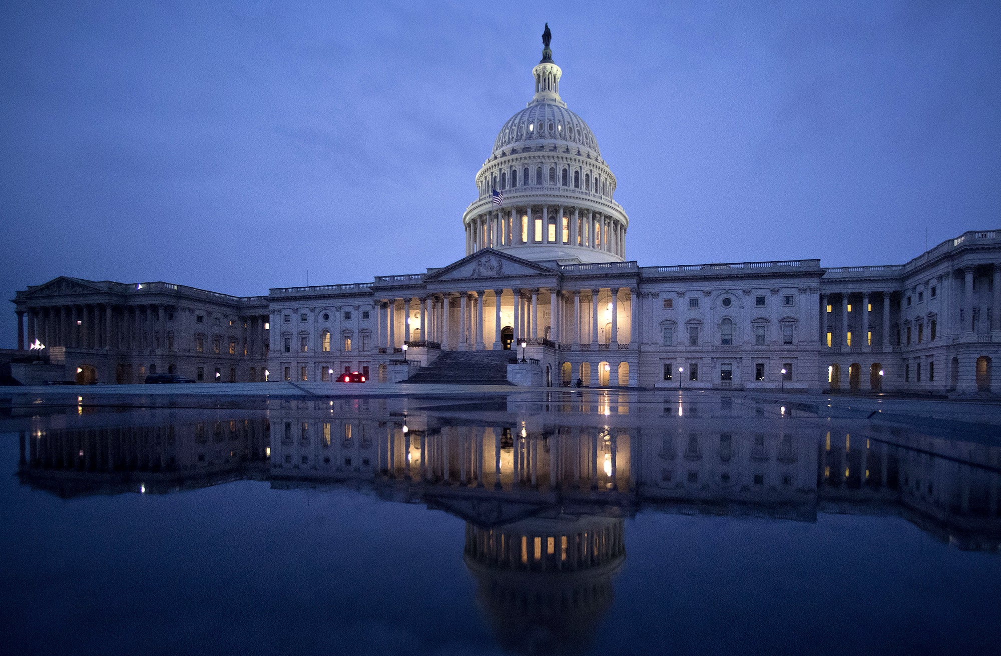 A wide photo of most of the U.S. Capitol building under a dark blue, cloudy sky. The building, with lights on, is reflected in water in the foreground.
